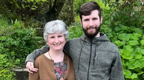 Anne Thompson Anne and Will Thompson stand in a garden with trees behind them. He has his arm around her shoulder and both are smiling.