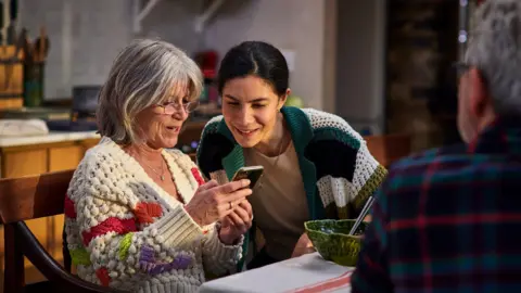 Getty Images Mother and adult daughter sit at a table looking at a phone, with an older man in the foreground and a bowl on the table.