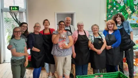 Silver Road Community Centre A group of people look into the camera and smile. Most of them are wearing aprons. They are stood inside a hall, with white walls behind them. In front there is a green plastic crate. 