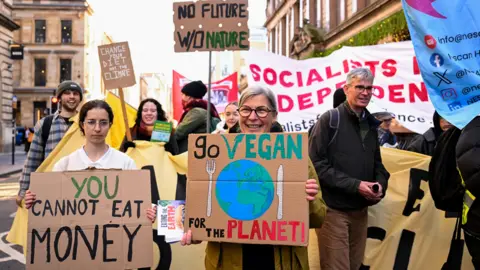 Reuters A group of climate protesters march down a city street holding handmade signs. At the front, a woman wearing glasses smiles while holding a cardboard sign that reads “Go Vegan for the Planet!” with a drawing of the Earth. Beside her, another protester holds a sign saying “You cannot eat money.” Behind them, more people carry banners and placards with messages such as “Change your diet, not the climate” and “No future without nature.” The crowd appears peaceful and engaged, with buildings lining the street in the background.