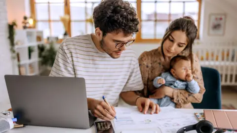 Getty Images A young couple with a baby sit at a desk looking at a laptop computer and a number of paper bills